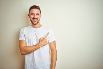 Young handsome man wearing casual white t-shirt over isolated background cheerful with a smile on face pointing with hand and finger up to the side with happy and natural expression