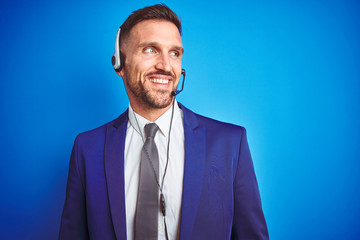 Young handsome operator man wearing call center headset over blue isolated background looking away to side with smile on face, natural expression. Laughing confident.