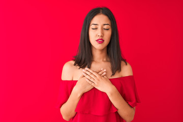Young beautiful chinese woman wearing casual t-shirt standing over isolated red background smiling with hands on chest with closed eyes and grateful gesture on face. Health concept.