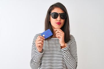 Young chinese woman wearing sunglasses holding credit card over isolated white background serious...