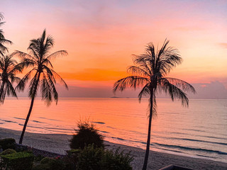Fototapeta na wymiar A picturesque tropical crimson red coloured coastal sunrise seascape with palm trees and ocean water reflection. Thailand.