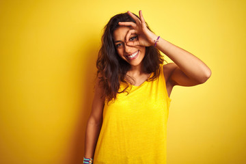 Young beautiful woman wearing t-shirt standing over isolated yellow background doing ok gesture with hand smiling, eye looking through fingers with happy face.