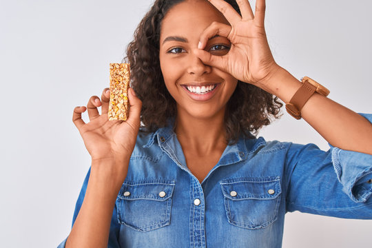 Young brazilian woman eating granola bar standing over isolated white background with happy face smiling doing ok sign with hand on eye looking through fingers