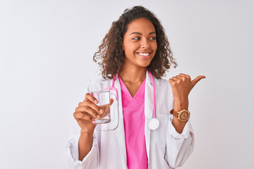 Young brazilian doctor woman holding glass of water standing over isolated white background pointing and showing with thumb up to the side with happy face smiling