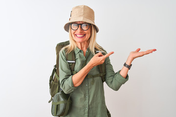Middle age hiker woman wearing backpack hat canteen glasses over isolated white background amazed and smiling to the camera while presenting with hand and pointing with finger.