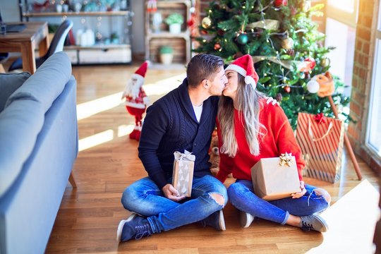 Young beautiful couple smiling happy and confident. Sitting on the floor holding gifts. Hugging and kissing around christmas tree at home