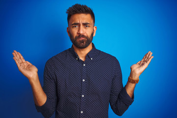 Young indian elegant man wearing shirt standing over isolated blue background clueless and confused...