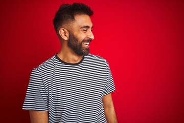 Young handsome indian man wearing navy striped t-shirt over isolated red background looking away to side with smile on face, natural expression. Laughing confident.