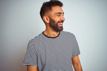 Young indian man wearing black striped t-shirt standing over isolated white background looking away to side with smile on face, natural expression. Laughing confident.