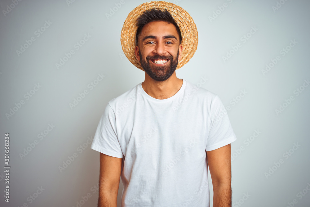 Wall mural young indian man on holiday wearing summer hat standing over isolated white background with a happy 