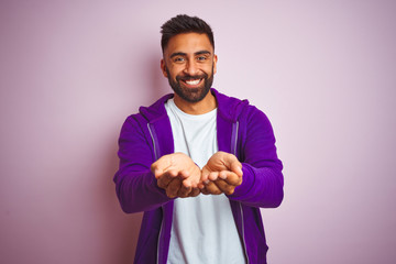 Young indian man wearing purple sweatshirt standing over isolated pink background Smiling with hands palms together receiving or giving gesture. Hold and protection