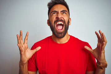 Young indian man wearing red t-shirt over isolated white background crazy and mad shouting and...