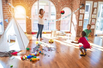 Young beautiful teacher and toddlers playing basketball around lots of toys at kindergarten