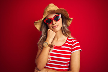 Young beautiful woman wearing sunglasses and summer hat over red isolated background with hand on chin thinking about question, pensive expression. Smiling with thoughtful face. Doubt concept.