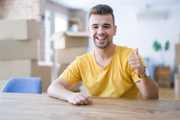 Young man sitting on the table with cardboard boxes behind him moving to new home doing happy thumbs up gesture with hand. Approving expression looking at the camera with showing success.
