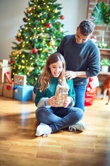 Young beautiful couple smiling happy and confident. Man surprise woman with gift around christmas tree at home