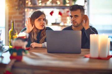 Young beautiful couple sitting using laptop around christmas decoration at home smiling with hand over ear listening an hearing to rumor or gossip. Deafness concept.