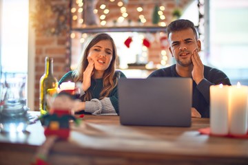 Young beautiful couple sitting using laptop around christmas decoration at home touching mouth with hand with painful expression because of toothache or dental illness on teeth. Dentist concept.