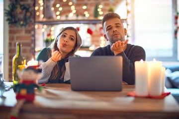 Young beautiful couple sitting using laptop around christmas decoration at home looking at the camera blowing a kiss with hand on air being lovely and sexy. Love expression.