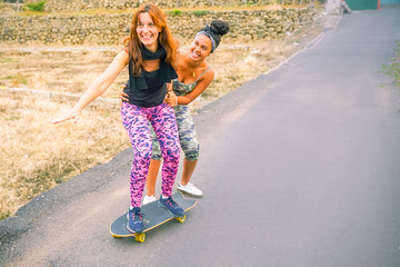 Young mother practising on skateboard in the street. Daughter helping mum on skateboard. Mum learning to ride skateboard as Daughter teaches her in the suburb street having fun - Image