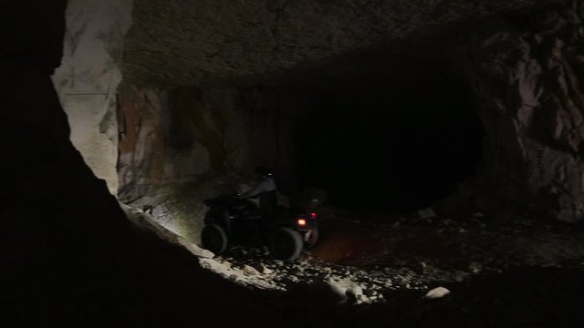 Several extreme quadrocycles switched on headlights and with men wearing helmets are actively riding and moving around inside of huge empty dark cave.