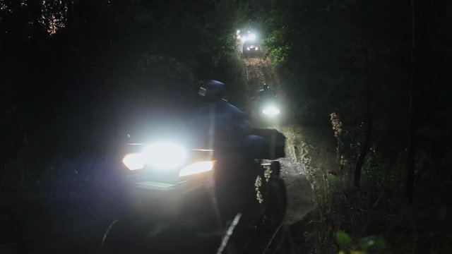 Row of several extreme quadrocycles switched on headlights and with men wearing helmets are actively riding and moving around inside of huge empty dark cave.