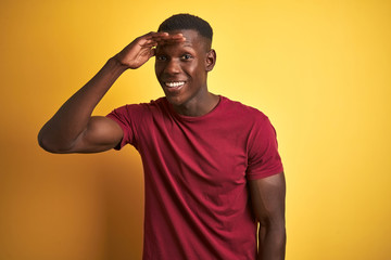 Young african american man wearing red t-shirt standing over isolated yellow background very happy and smiling looking far away with hand over head. Searching concept.