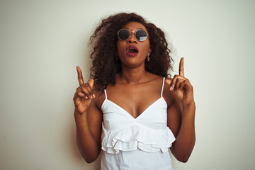 Young african american woman wearing t-shirt and sunglasses over isolated white background amazed and surprised looking up and pointing with fingers and raised arms.