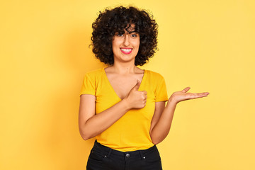 Young arab woman with curly hair wearing t-shirt standing over isolated yellow background Showing palm hand and doing ok gesture with thumbs up, smiling happy and cheerful