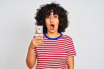Young arab woman with curly hair holding dollars standing over isolated white background scared in shock with a surprise face, afraid and excited with fear expression