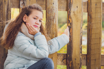 Beautiful girl in scarf and blue down jacket is squatting holding wooden fence in the village on an autumn cool day.
