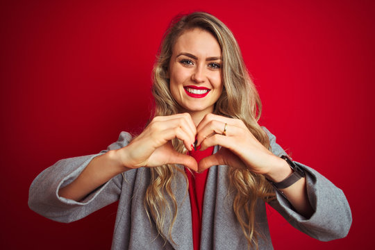 Young beautiful business woman wearing elegant jacket standing over red isolated background smiling in love doing heart symbol shape with hands. Romantic concept.