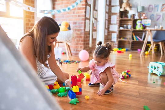 Young beautiful teacher and toddler sitting on the floor playing with building blocks toy at kindergarten