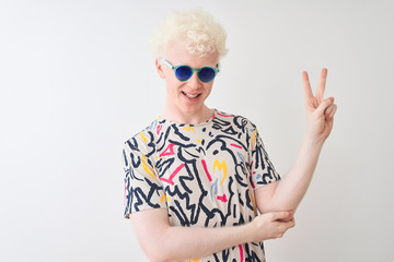Young albino blond man wearing colorful t-shirt and sunglasses over isolated red background smiling with happy face winking at the camera doing victory sign. Number two.