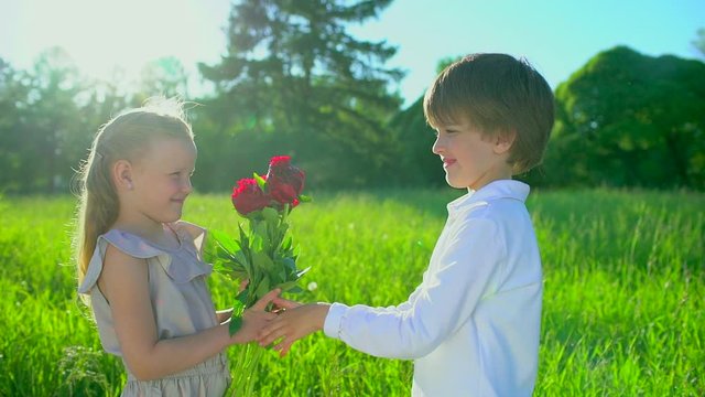 ittle cute boy giving to beautiful girl bouquet flowers peonies outdoors at sunset. first love, friendship, childhood, Funny kids, child. lovely childrens couple in nature, summer, slow-motion