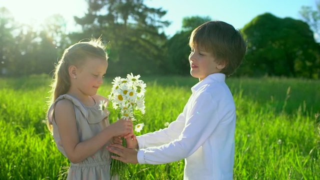 ittle cute boy giving to beautiful girl bouquet flowers chamomiles outdoors at sunset. first love, friendship, childhood, Funny kids, child. lovely childrens couple in nature, summer, slow-motion