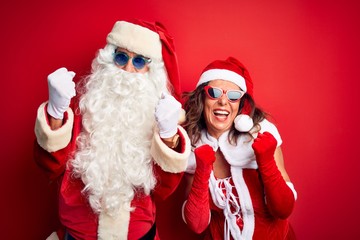 Middle age couple wearing Santa costume and sunglasses over isolated red background very happy and excited doing winner gesture with arms raised, smiling and screaming for success