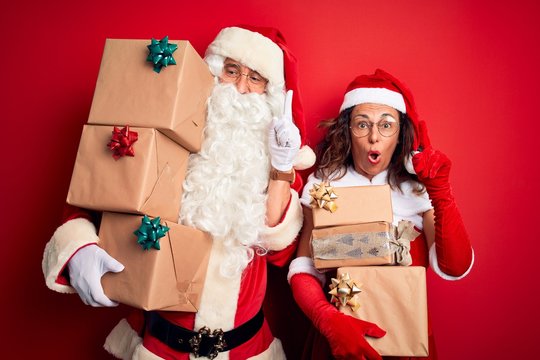 Middle age couple wearing Santa costume holding tower of gifts over isolated red background pointing finger up with successful idea. Exited and happy. Number one.