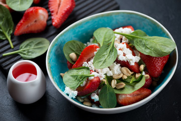 Salad with spinach leaves, fresh strawberries, cottage cheese and nuts served in a turquoise bowl, studio shot over black stone surface