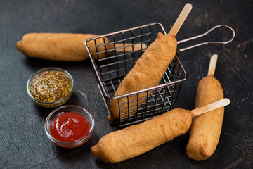 Deep-fried sausages covered in a dough and served with ketchup and mustard. Studio shot over dark brown stone background
