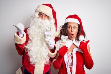 Senior couple wearing Santa Claus costume holding dollars over isolated white background Surprised pointing with hand finger to the side, open mouth amazed expression.