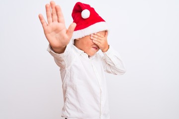 Beautiful kid boy wearing Christmas Santa hat standing over isolated white background covering eyes with hands and doing stop gesture with sad and fear expression. Embarrassed and negative concept.