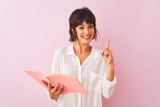 Young beautiful teacher woman reading book standing over isolated pink background surprised with an idea or question pointing finger with happy face, number one