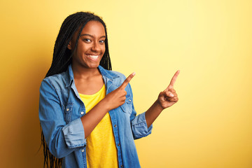 Young african american woman wearing denim shirt standing over isolated yellow background smiling and looking at the camera pointing with two hands and fingers to the side.