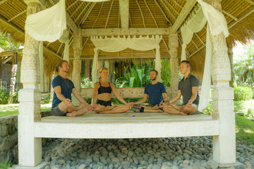 Obraz na płótnie Canvas happy group of young hipster American friends enjoying Asian yoga retreat together sitting on lotus position meditating at outdoors hut in Bali in mind body harmony