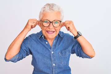 Senior grey-haired woman wearing denim shirt and glasses over isolated white background Smiling pulling ears with fingers, funny gesture. Audition problem