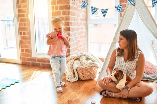 Beautiful teacher sitting on the floor getting angry with blond toddler girl outside tipi at kindergarten