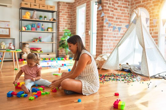 Beautiful teacher and blond toddler girl building tower using plastic blocks at kindergarten