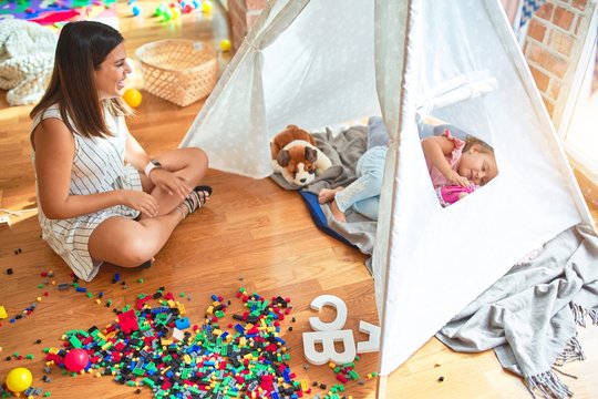 Beautiful teacher and blond toddler girl playing with dolls inside tipi at kindergarten