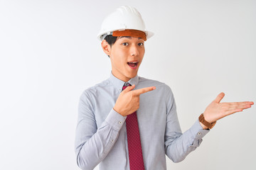 Chinese architect man wearing tie and helmet standing over isolated white background amazed and smiling to the camera while presenting with hand and pointing with finger.